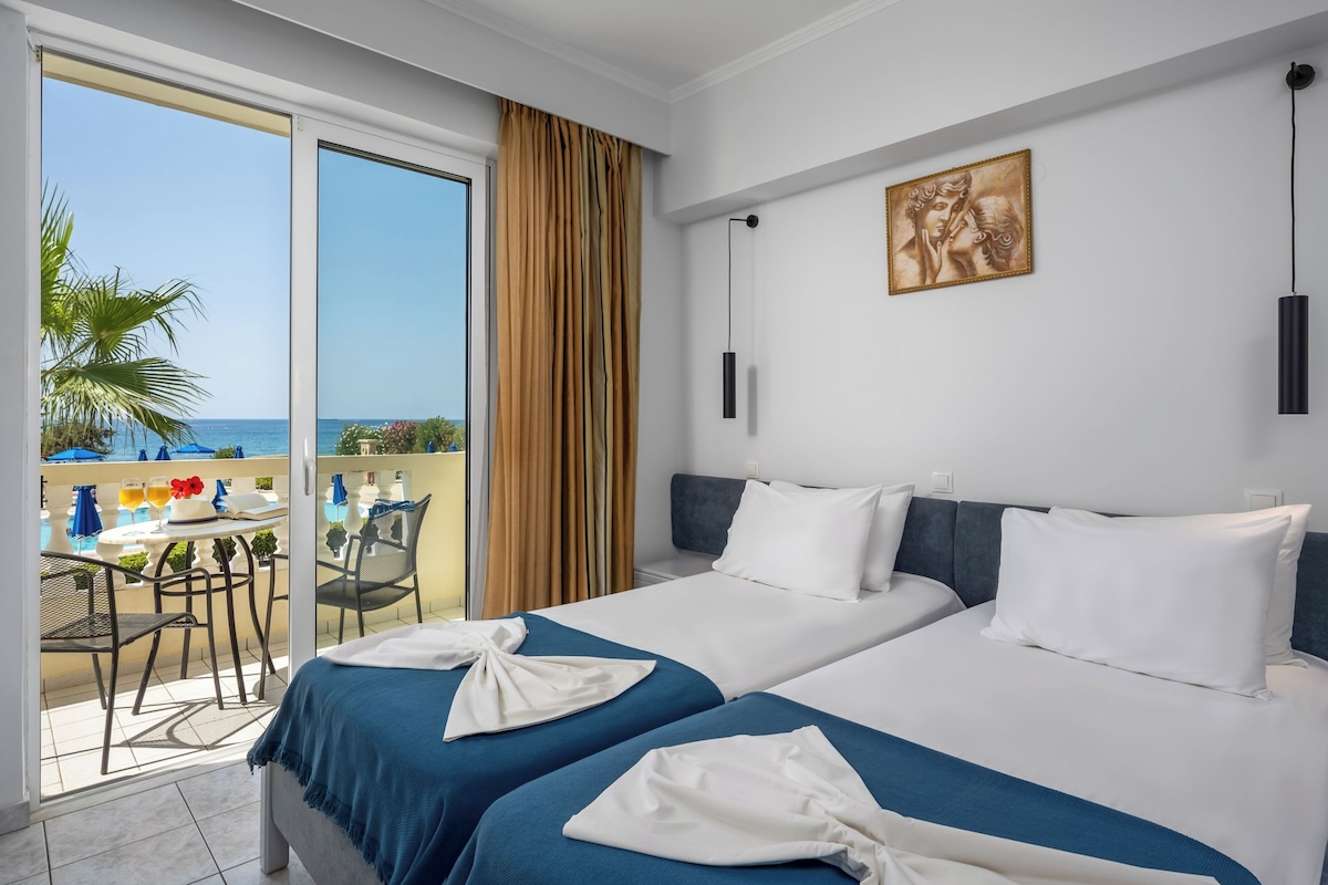 Quarto de hotel com duas camas, varanda com vista para o mar e área de refeições com mobiliário para área externa.