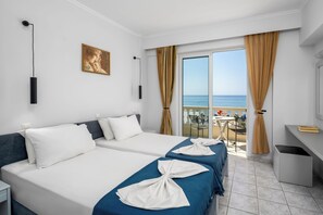 Quarto de hotel com duas camas, uma mesa, uma cadeira, uma janela com vista para o mar e um quadro na parede.