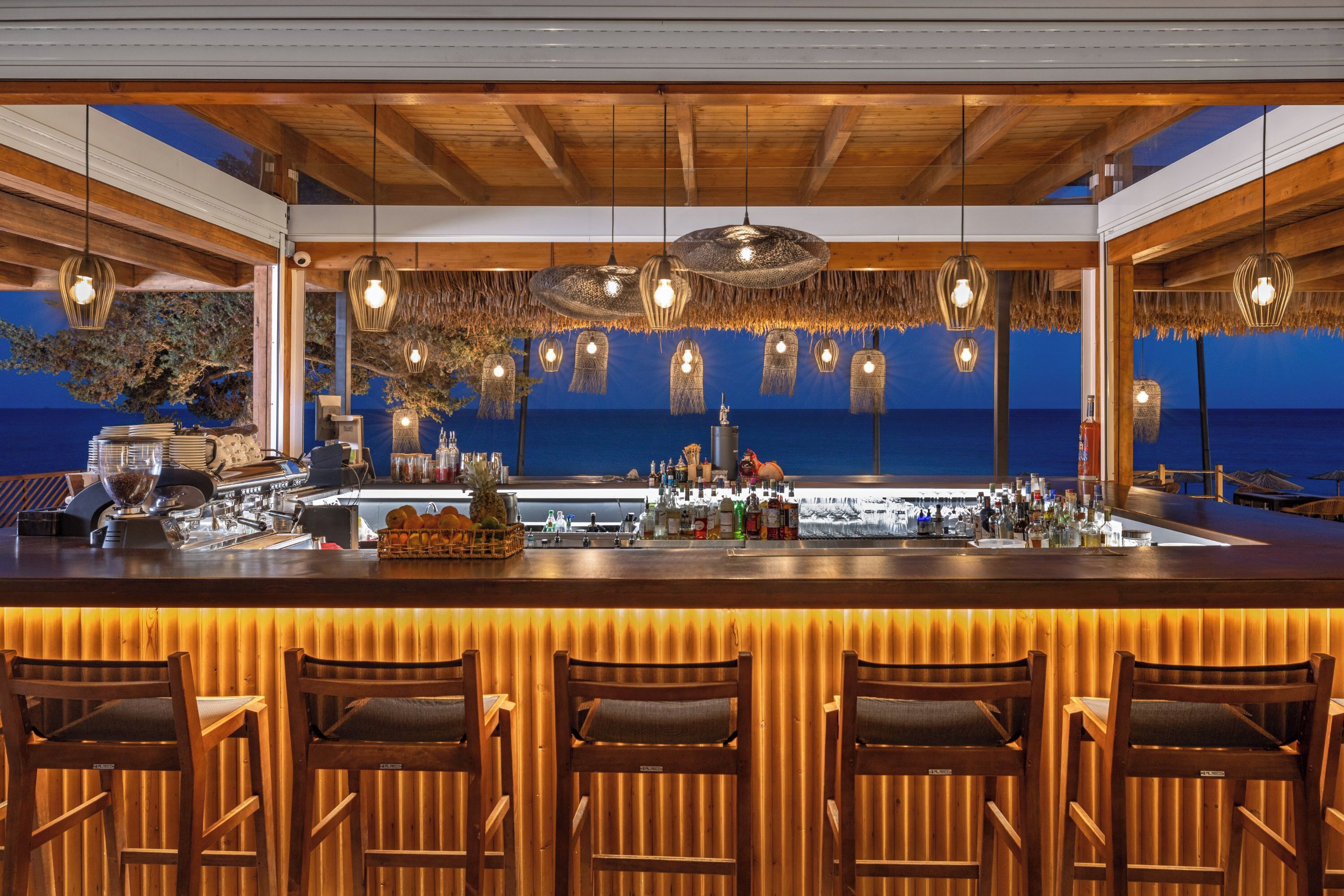 A bar with wooden bar stools, a counter with various bottles, and a view of the sea.