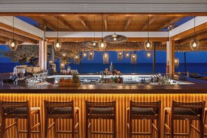 Um bar com banquetas de madeira, um balcão com diversos garrafas e vista para o mar.