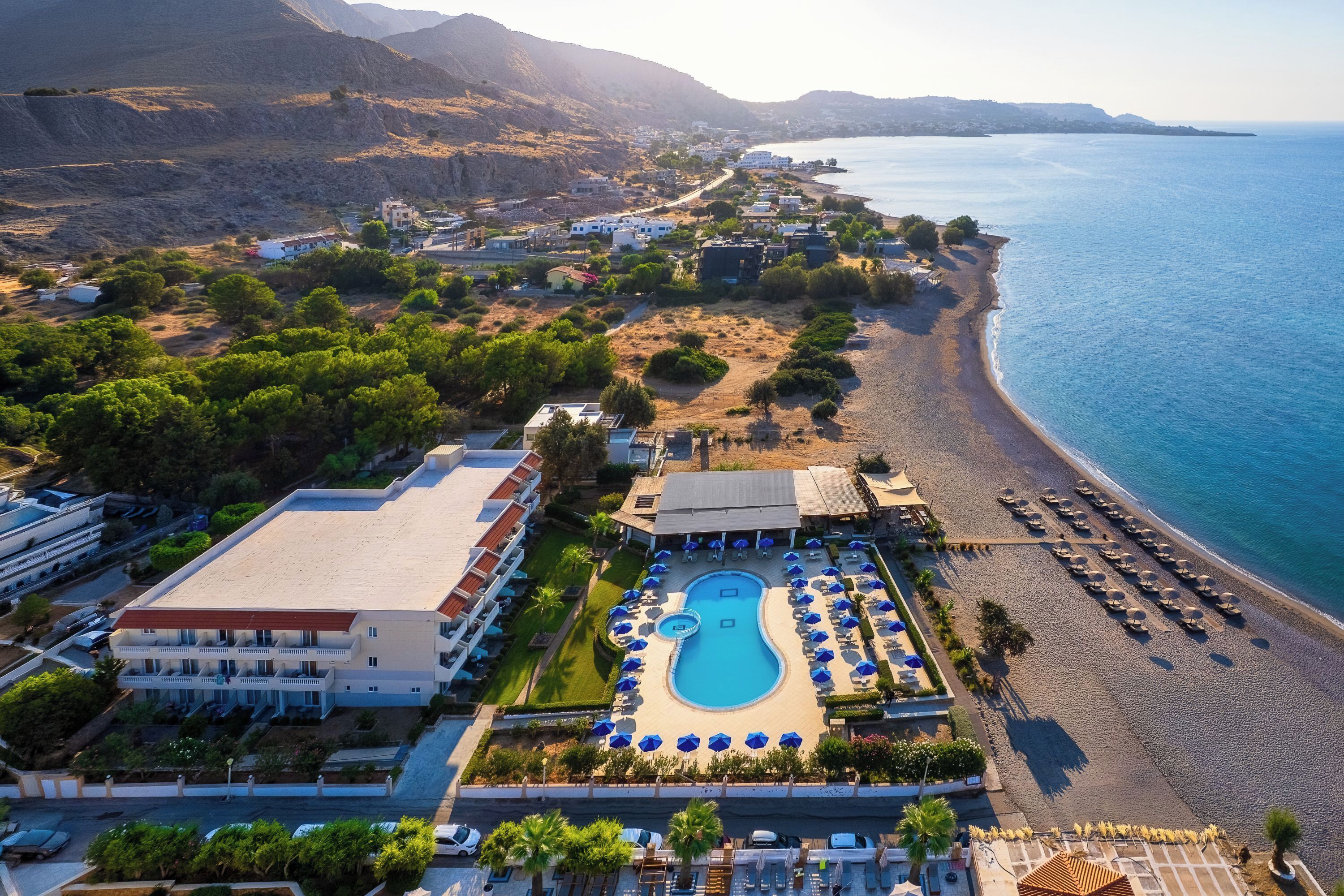 An aerial view of a resort with a swimming pool, beach, and mountainous backdrop.