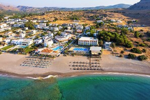 Vista aérea de um resort à beira-mar, com vários edifícios, uma praia com espreguiçadeiras e águas turquesa cristalinas.
