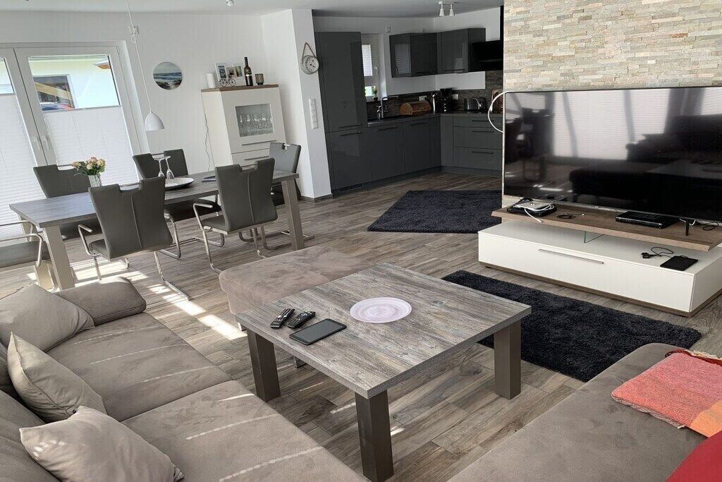 Una sala de estar moderna con un sofá, una mesa de centro y un televisor. Se observa un comedor con sillas y una mesa. Se ve la cocina al fondo.
