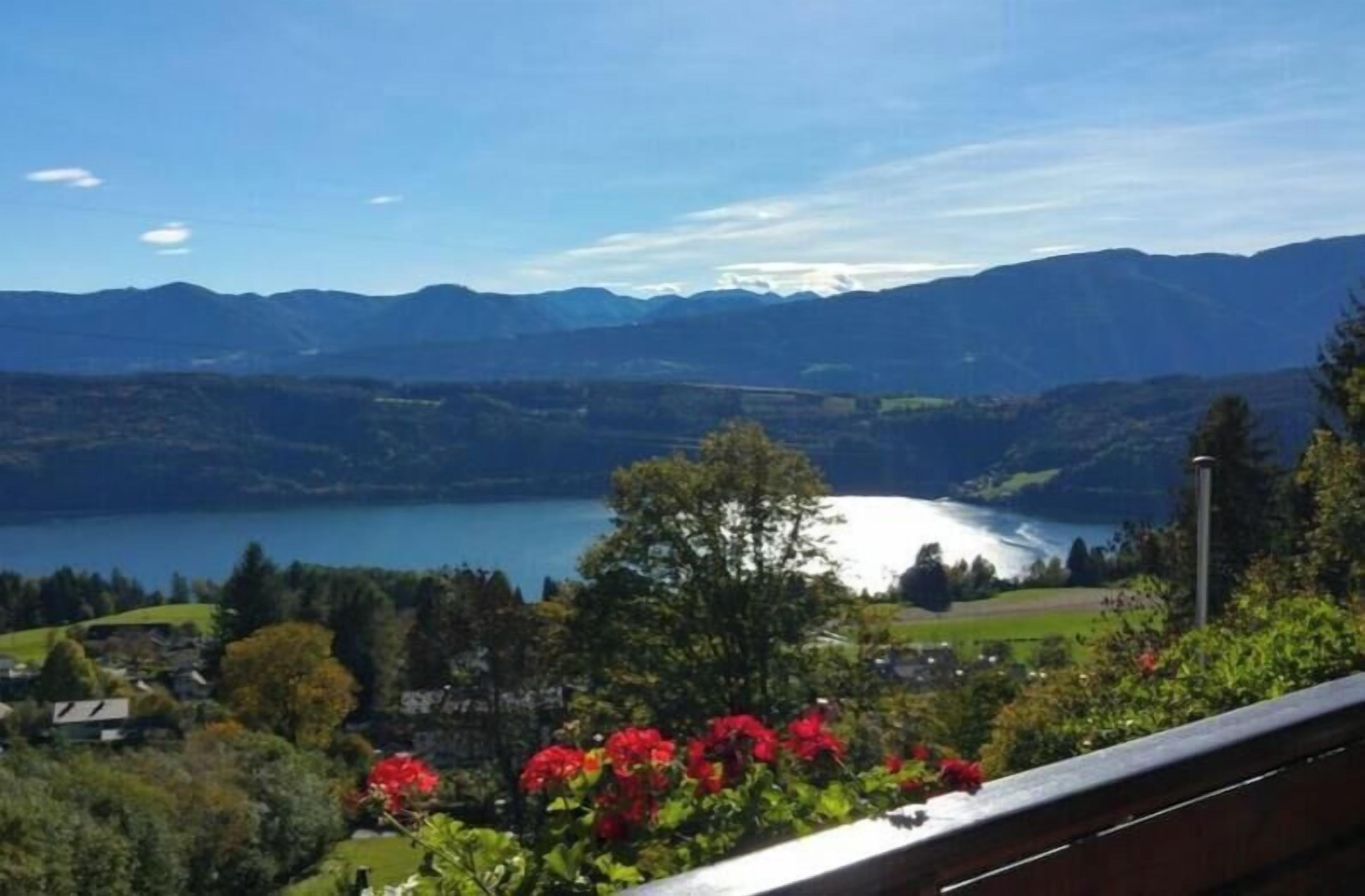 Une vue pittoresque d’un lac, avec des montagnes en arrière-plan, un balcon orné de fleurs rouges et un ciel dégagé.