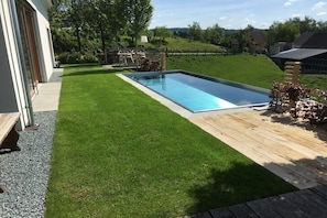 Una piscina rettangolare con un bordo in legno, circondata da un prato curato e con vista su case in lontananza.