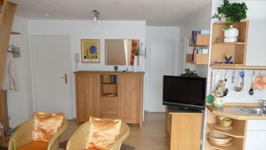 A compact living space with a kitchenette, a television, wicker furniture, and a small dining area. - Vrbo Property (Ostseebad Prerow)