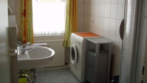 A compact bathroom with a sink, a washing machine, and a window with curtains. - Vrbo Property (Ostseebad Prerow)