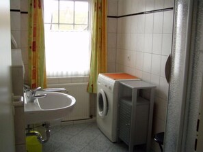 Een kleine badkamer met een wastafel, een wasmachine en een raam met gordijnen.