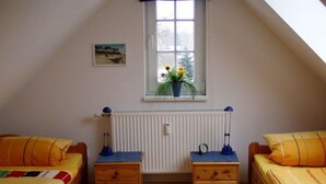 A room with two beds, a window with plants, a radiator, and a small table with a lamp. - Vrbo Property (Ostseebad Prerow)
