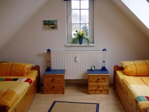 Een kamer met twee bedden, een raam met planten, een radiator en een klein tafeltje met een lamp.