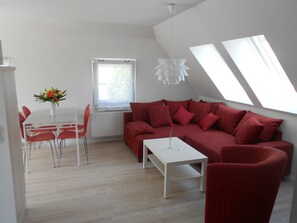 Ein modernes Wohnzimmer mit einem roten Sofa, einem weißen Couchtisch und roten Sesseln. Es gibt einen weißen Heizkörper, ein Fenster mit Blick und eine Deckenleuchte.