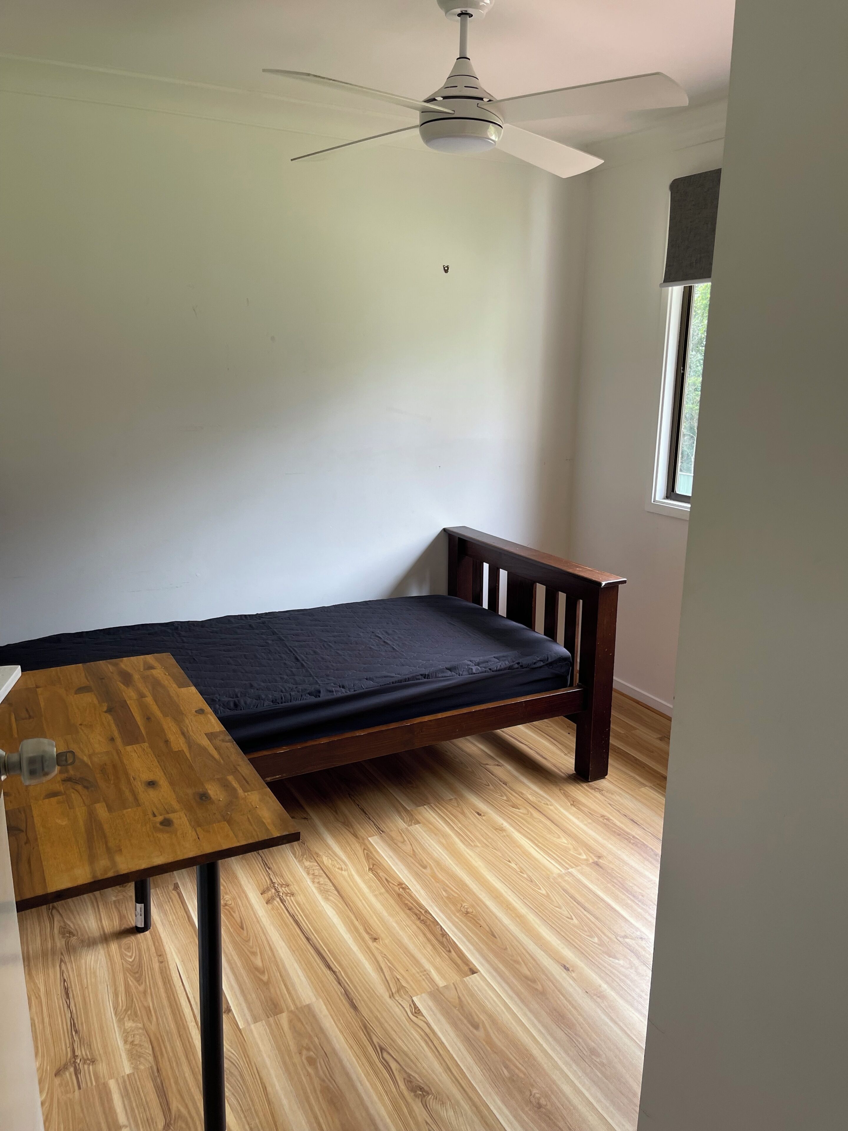 Ein kleines Zimmer mit einem Einzelbett, einem Holztisch, einem Deckenventilator und einem Fenster.