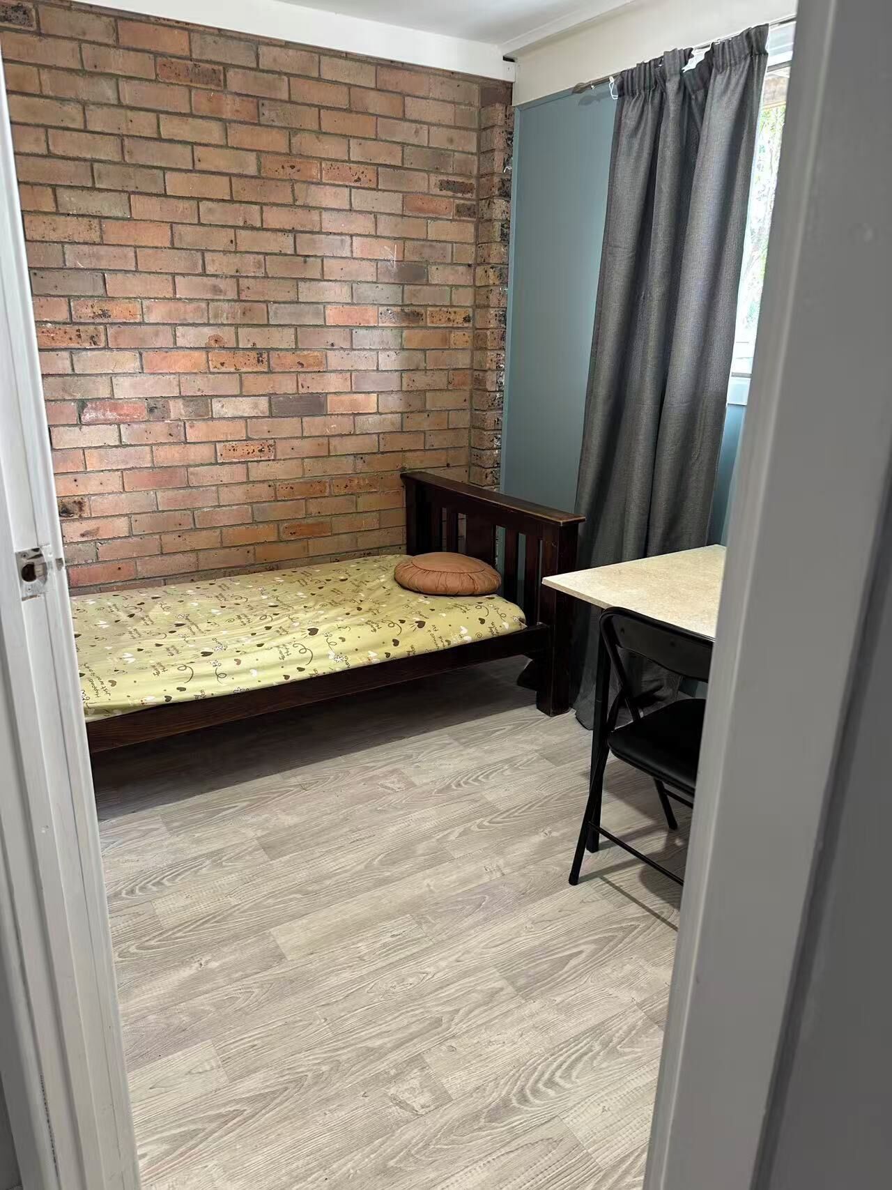Una habitación pequeña con pared de ladrillo, cama individual con colcha estampada, mesa de madera y una silla negra.