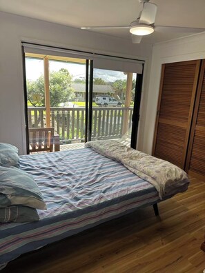 A bedroom with a bed, a ceiling fan, a wooden floor, and a view of a residential area through a sliding door. - Tranquil Waters Edge Escape - 4BR Waterfront Home - family holiday retreat (Sanctuary Point)