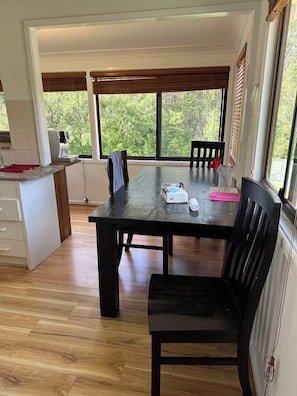 A dining area with a wooden table and chairs, a kitchen area with a coffee machine, and large windows with a view of greenery. - Tranquil Waters Edge Escape - 4BR Waterfront Home - family holiday retreat (Sanctuary Point)