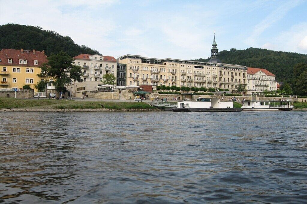 A riverside building with a prominent tower, a boat docked in front, and a hilly backdrop.