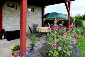 A stone building with a covered patio area featuring a table and chairs, surrounded by various potted plants and flowers. - Holiday apartment with 1 bedroom (Rathmannsdorf)