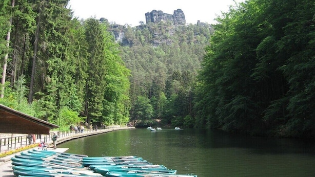 Vista para um lago com barcos atracados, um deck de madeira e uma colina arborizada com formações rochosas ao fundo.
