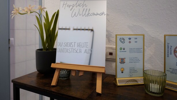 Een houten tafel met een bord waarop 'Herzlich Willkommen' staat en een bloempot met plant.
