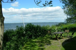 Eine grüne Fläche mit Tisch und Stühlen, einer Bank und Blick auf ein Gewässer mit einem Segelboot in der Ferne.