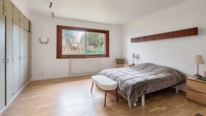 Ein Schlafzimmer mit Bett, einer Bank, einem Nachttisch, einer Lampe und einem Fenster mit Blick auf Grünflächen.