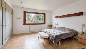 Une chambre à coucher comprenant un lit, un banc, une table de chevet, une lampe et une fenêtre donnant sur un paysage verdoyant.