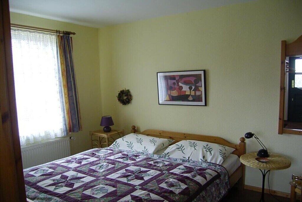 Une chambre à coucher avec un lit, une petite table, une lampe, un tableau encadré au mur et une fenêtre avec des rideaux.