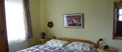 Une chambre à coucher avec un lit, une petite table, une lampe, un tableau encadré au mur et une fenêtre avec des rideaux.