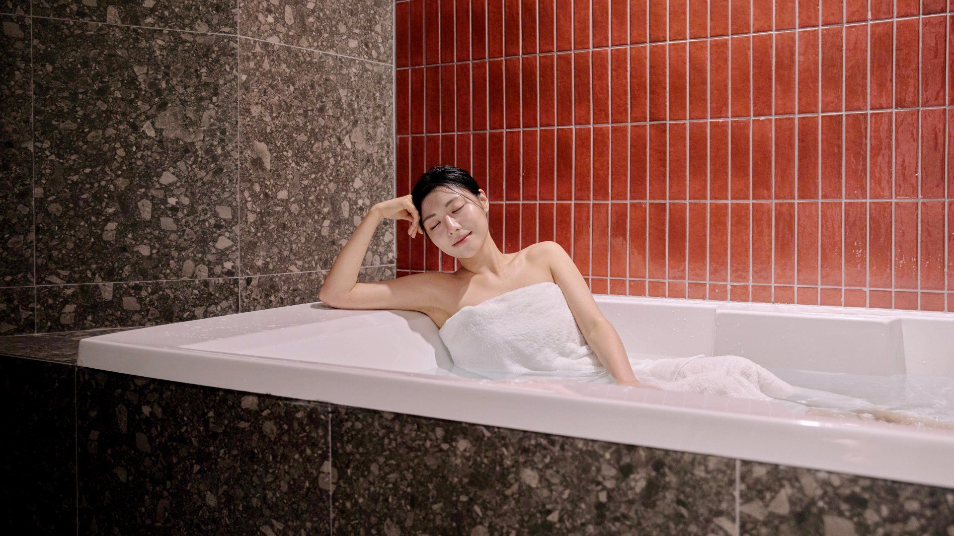 A person in a bathtub with a towel wrapped around their body, set against a background of red tiles and a speckled wall.
