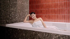 A person in a bathtub with a towel wrapped around their body, set against a background of red tiles and a speckled wall. - Muonjae by Anook Suwon Heanggung (Suwon)