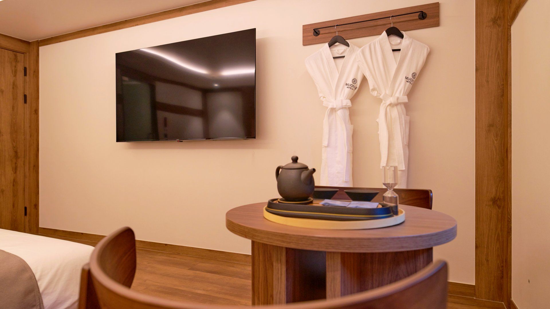 A hotel room with a flat-screen TV mounted on the wall, a wooden sideboard with a teapot and a tray, and two white robes hanging on a wooden rack.