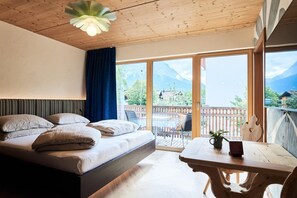 A bedroom with a bed, a table, and a view of mountains through large windows. - Vrbo Property (Lermoos)