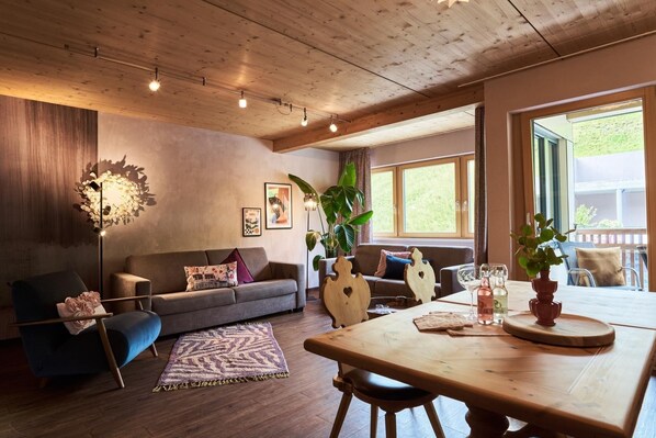 A cozy living room with a sofa, a dining table, and a large window. - Vrbo Property (Lermoos)