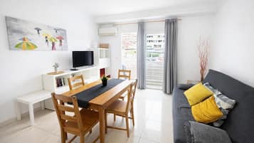 Una sala moderna con comedor, un sofá con cojines, un televisor de pantalla plana, una estantería y una ventana con cortinas.