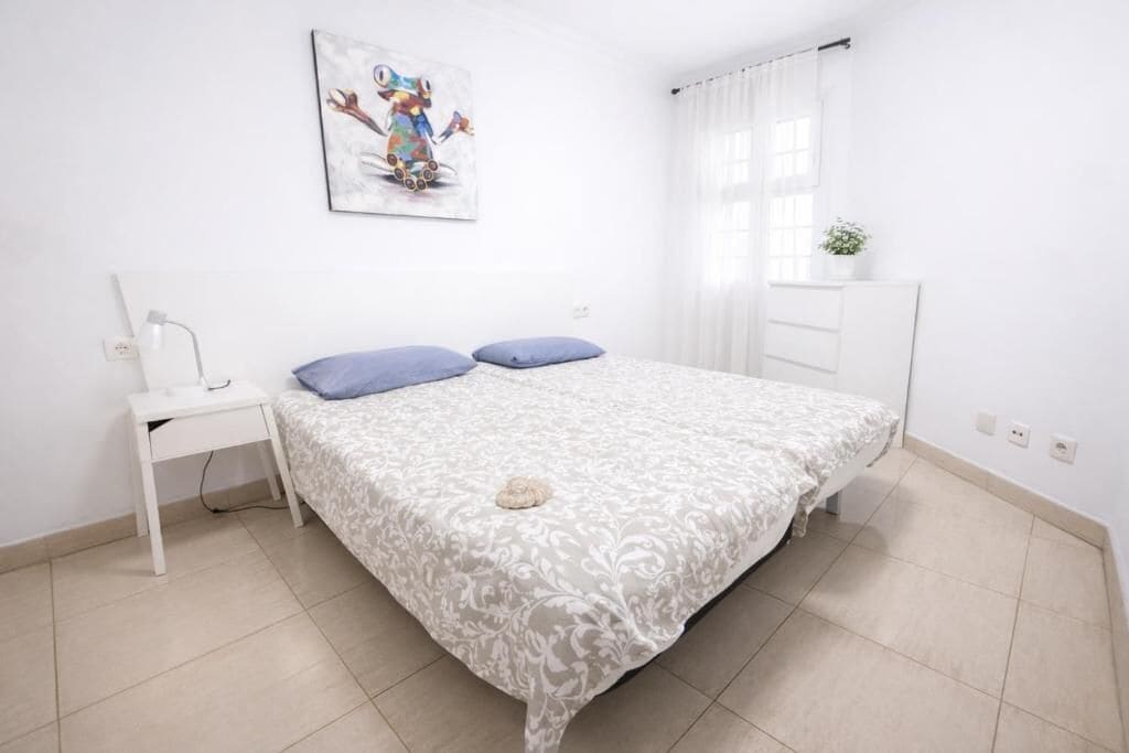 Un dormitorio con cama, mesita de noche, lámpara de pared, ventana con cortinas y una planta pequeña en maceta.