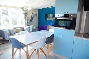 A modern kitchen with a built-in oven and microwave, a dining area with a white table and chairs, and a living space with a sofa and a TV. - Holiday home with 2 bedrooms (Westende-Dorp)