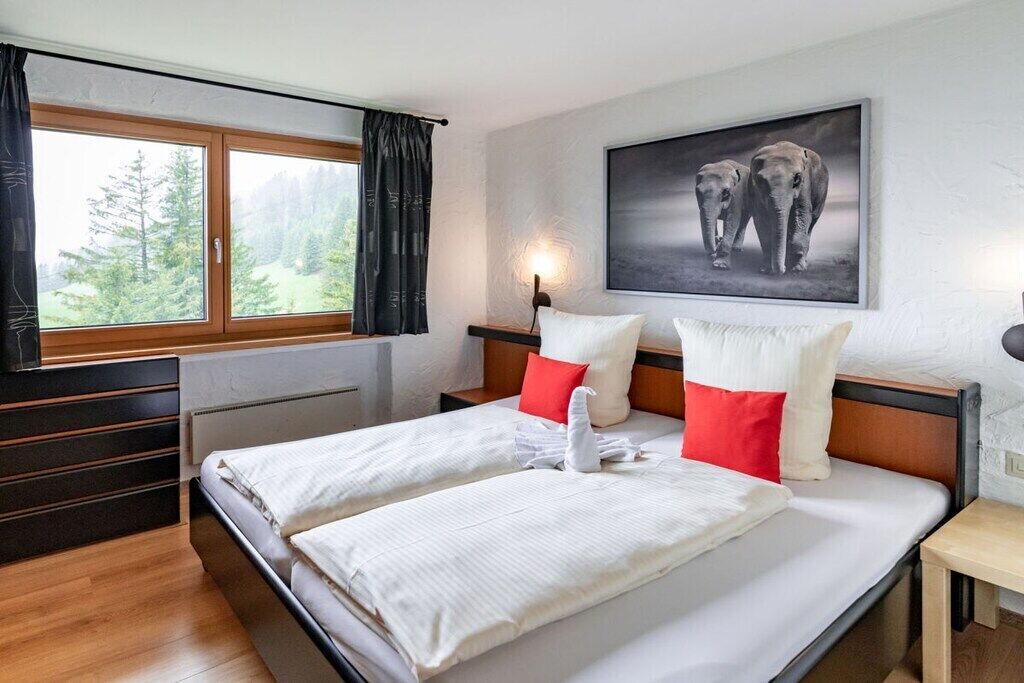 A hotel room with a large bed, two pillows, a nightstand, a lamp, and a window with curtains.