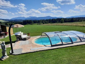 En swimmingpool med en glasvägg runt om, omgiven av en uteplats med utemöbler, en staty och utsikt över ett bergigt landskap.