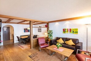 Ein Zimmer mit einem Holztisch, einer Couch mit Kissen, einem roten Sessel und einer Pflanze.