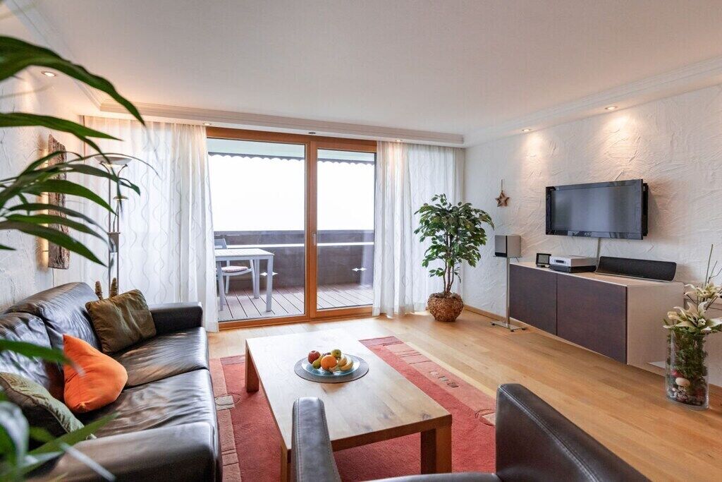 Un salon moderne avec un canapé en cuir, une table basse en bois sur laquelle sont disposés des fruits, un téléviseur fixé au mur et une porte-fenêtre coulissante donnant sur un espace extérieur.