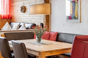 Et spiseområde med et træbord, stole, en sofa med mønstrede hynder og en vase med røde blomster.