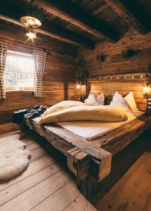 A rustic wooden cabin bedroom with a bed, pillows, a fur rug, and a window with curtains. - Vrbo Property (Saalbach)