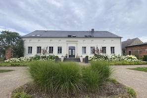 Ein zweistöckiges Gebäude mit einer symmetrischen Fassade, einem zentralen Eingang mit Stufen und einem Garten mit hohem Gras und weißen Blüten davor.