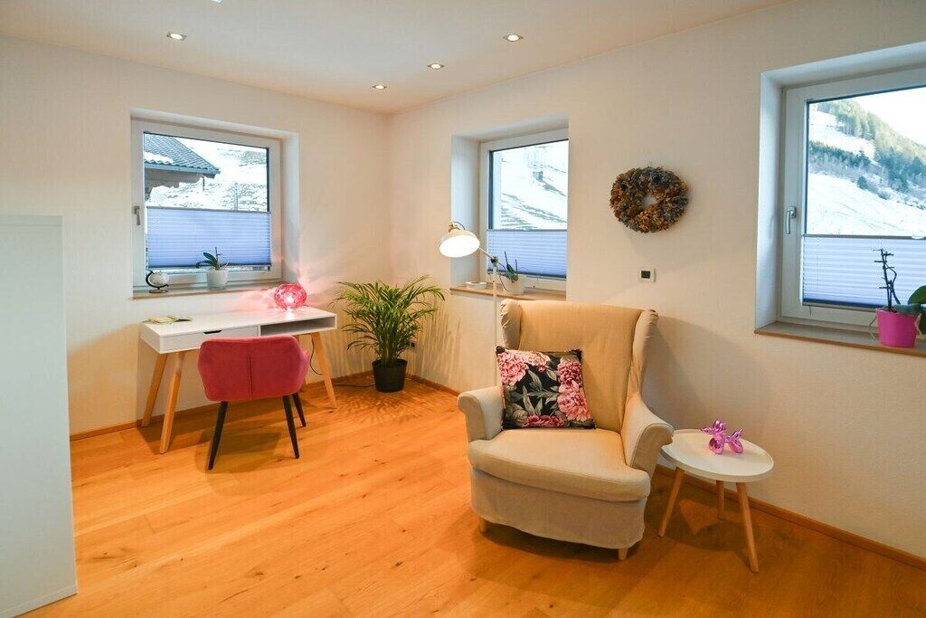 A room with a desk, chair, armchair, small table, potted plants, and windows with views of a snowy landscape.