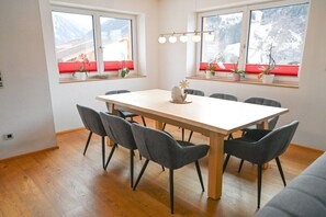 A dining area with a wooden table and six chairs, a window with red blinds, and a view of mountains. - Holiday home in Rauris with swimming pool (Rauris)