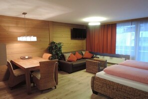 Una camera d'albergo con parete in legno, una TV montata sopra un divano, un tavolo da pranzo con sedie e un letto con copriletto a quadri.