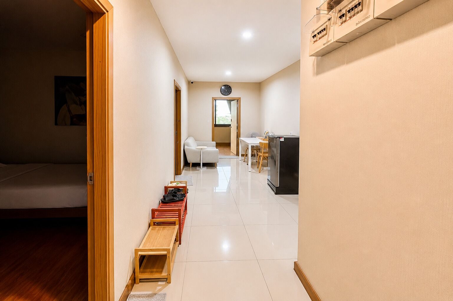 A hotel room with a bed, a wooden bench, a small table, a chair, and a kitchenette area.