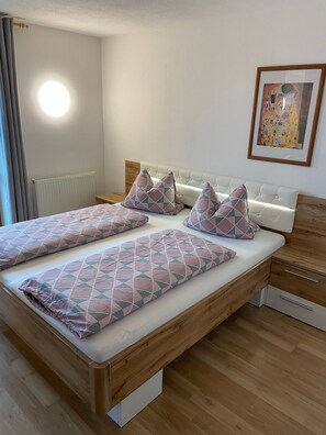 A double bed with a geometric patterned bedspread and pillows, a wooden headboard, a bedside table, a drawer unit, a radiator, and a framed artwork on the wall. - Vrbo Property (Ehrwald)