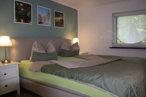 A neatly made bed with pillows and a blanket, a bedside table with a lamp, and a window with a view of trees. - Holiday home with 2 bedrooms (Burg (Spreewald))