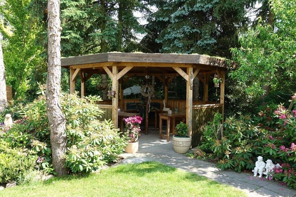 A wooden gazebo with a table and chairs, surrounded by lush greenery and potted plants.